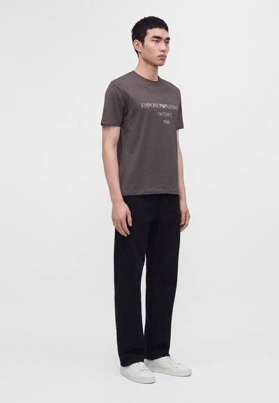 Νέος άνδρας που στέκεται, φορώντας γκρι t-shirt Emporio Armani, μαύρο παντελόνι και λευκά αθλητικά παπούτσια, μπροστά σε απλό φωτεινό φόντο.