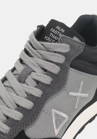 Scarpa sportiva grigia con tomaia in tessuto testurizzato, suola in gomma e accenti scuri a contrasto. Include lacci e logo sulla linguetta.
