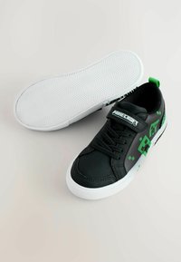 Zapatillas negras con superficie texturizada, suela blanca, acentos pixelados verdes y una correa con la marca "Minecraft".