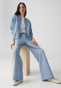 Veste bomber bleu clair avec zip sur le devant, associée à un pantalon large assorti. Texture lisse, design décontracté, portée avec des ballerines crème.