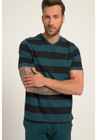 Stripet kortærmet skjorte i teal og sort, med V-hals-design og blød stof. Modellen står med armene over kors, så tatoveringsdetaljerne vises.