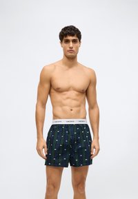 Bărbat muscular în picioare, purtând boxeri în carouri albastre și verzi cu logo-uri Lacoste albe și un elastic alb cu textul Lacoste.