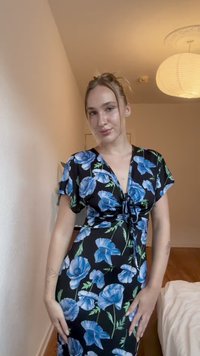 Vestido negro con mangas cortas, adornado con grandes patrones florales azules y un detalle de lazo en la parte delantera, fabricado en tela suave. El fondo es neutro.
