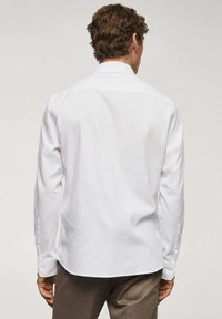 Chemise blanche à manches longues avec col pointu, texture lisse, poignets à boutons, et ourlet arrondi. Vue de dos.