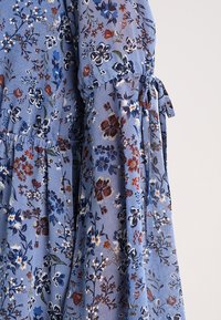 Vestido floral azul con un tejido ligero, mangas acampanadas y detalles con cordón en la cintura. Los patrones incluyen flores oscuras y claras.