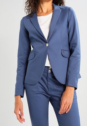 Kvinde iført en tætsiddende blå blazer med knappede lommer over en hvid top, kombineret med matchende blå skræddersyede bukser.