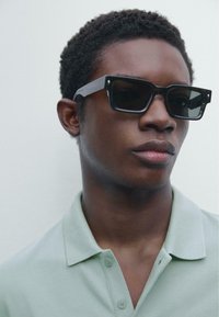 Svarta rektangulära solglasögon med tjocka bågar, prydda med silverdetaljer. Bärs tillsammans med en ljusgrön polo-tröja. Bakgrunden är neutral.