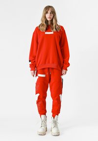 Sudadera roja de gran tamaño con una etiqueta blanca y pantalones rojos con patrones bordados en blanco, complementados con botas beige con cordones.