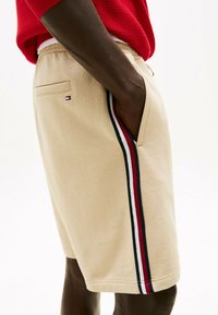 Pantaloni corti beige con strisce laterali rosse, bianche e blu e un piccolo logo sulla tasca posteriore, indossati con una maglietta rossa.