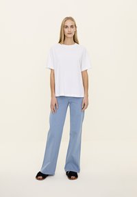 Bela majica s kratkimi rokavi, sproščenega kroja, v kombinaciji s svetlo modrimi širokimi hlačami. Model nosi črne krznene natikače. Preprost, sproščen outfit.