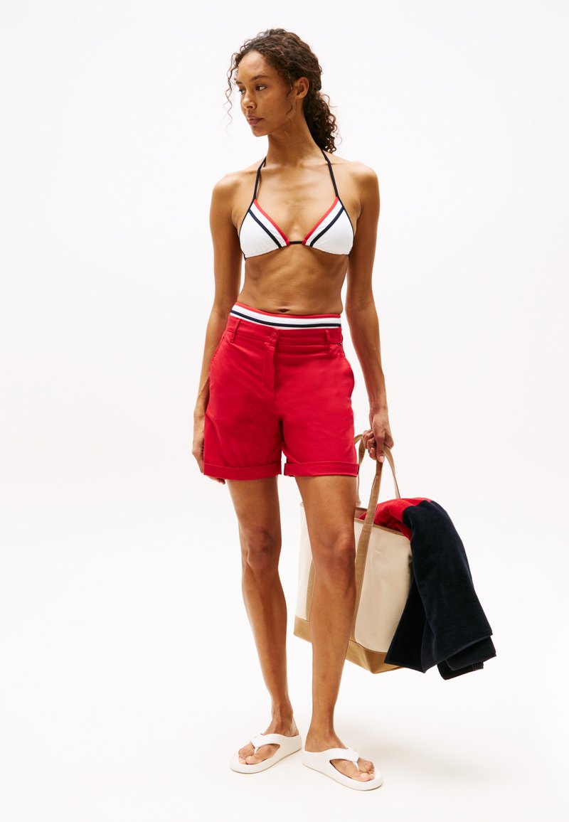 Pantaloni scurți roșii cu tivuri pliate, asortați cu un top de bikini alb și negru în formă de triunghi. Modelul ține o geantă de tip tote bej, plină cu îmbrăcăminte.