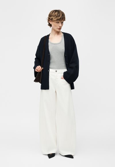 Femme portant un cardigan noir sur un débardeur gris, un pantalon blanc à jambes larges, des chaussures pointues noires, et tenant un sac à bandoulière noir.