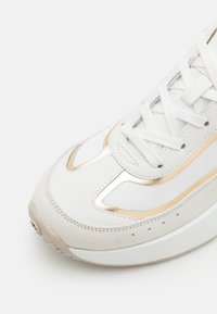 Baskets blanches présentant un mélange de cuir et de daim, des accents métalliques dorés, un design à lacets et une semelle texturée avec un bout arrondi.