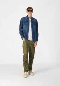 Denim jaka, zaļas kargo bikses, baltā t-krekls. Saulesbrilles, krēmveida apavi. Iezīmes: pogām sagatavoti kabatas, brīvs piegriezums, teksturēta auduma.