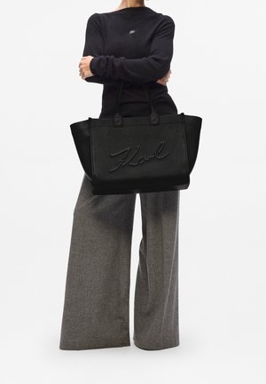 Personne portant un haut noir à manches longues et un pantalon large gris, tenant un grand sac fourre-tout noir texturé avec "Karl" embossé sur le devant.