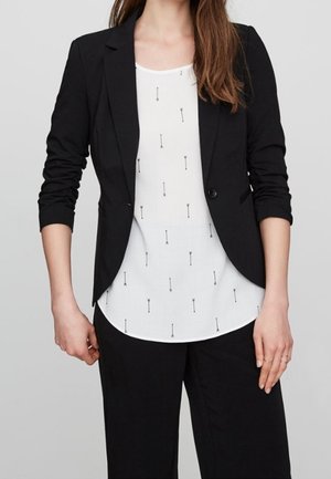 Femme portant un blazer noir à manches froncées sur un chemisier blanc avec un petit motif flèche noir, associé à un pantalon noir.