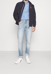 Męska ciemnogranatowa kurtka z zamkiem z przodu, jasnoniebieska koszula w kratę, jasne dżinsy i białe trampki z szarymi akcentami. Luźny krój.