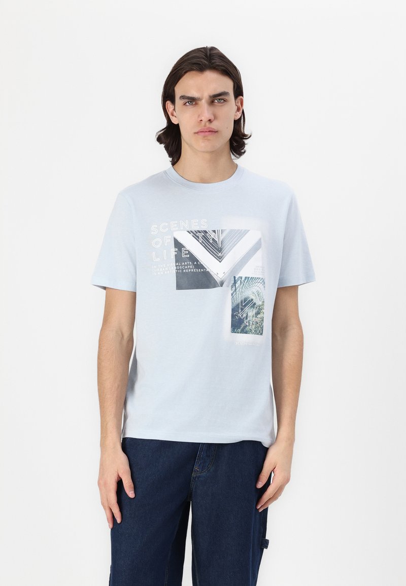 T-shirt en coton bleu clair avec un col rond, présentant des impressions graphiques multicolores et du texte dans un style collage sur le devant.