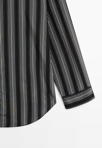 Manche de chemise noire et ourlet latéral avec fines rayures blanches et grises verticales, poignet avec rayures horizontales pour contraste.