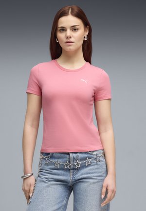 Young woman wearing a pink Puma t-shirt, light blue jeans, silver star chain belt, hoop earrings, and a bracelet against a gray background.