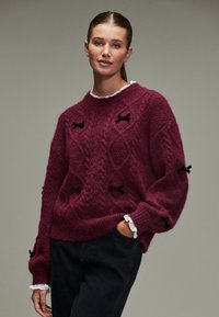 Bordeauxroter Strickpullover mit strukturiertem Muster und schwarzen Bandakzenten auf der Vorderseite. Lange, lockere Ärmel mit einem gerafften Saum.