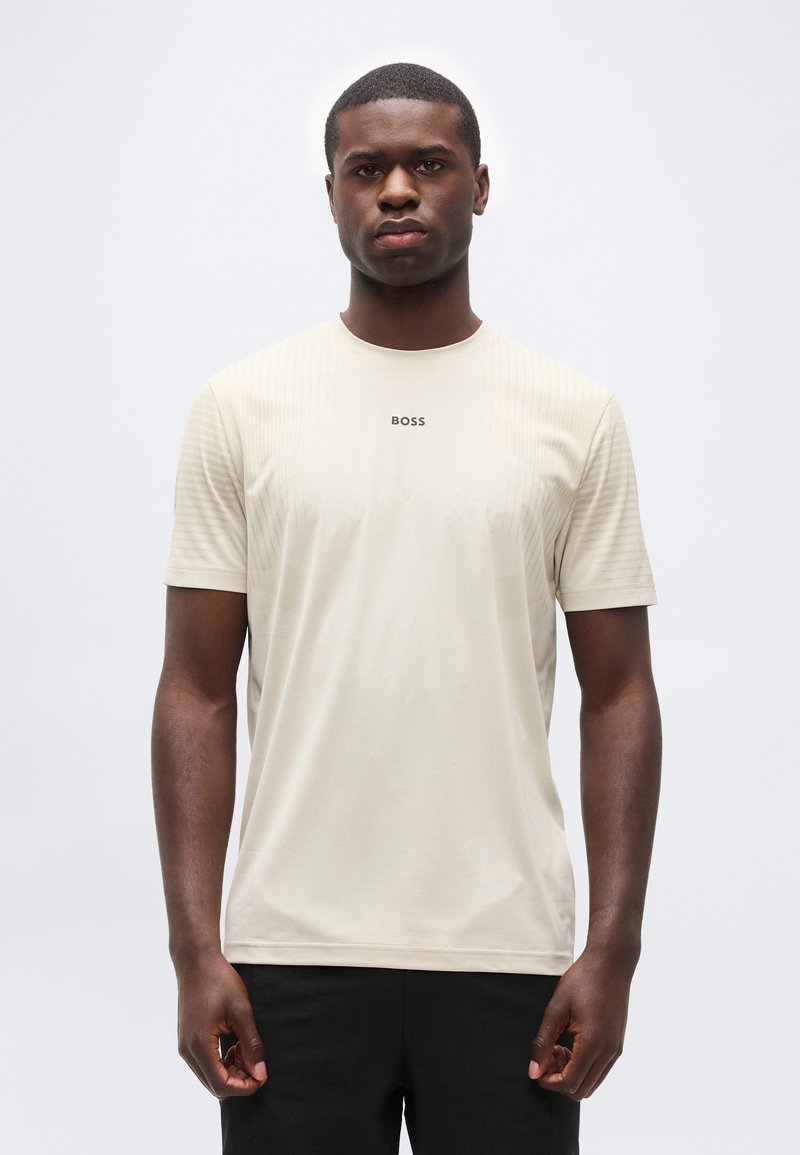 Νεαρός άνδρας που φοράει μια μπεζ κοντομάνικη μπλούζα BOSS, στέκεται μπροστά σε ένα απλό ανοιχτό γκρι φόντο με ουδέτερη έκφραση.