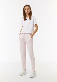 Weißes T-Shirt mit kurzen Ärmeln, kombiniert mit hellrosa Jogginghosen mit Kordelzug, die Seitentaschen und eine Logo-Stickerei aufweisen, abgerundet mit weißen Sneakers.