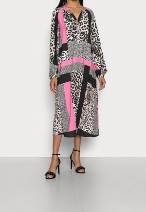 Vrouw draagt een midi-jurk met lange mouwen met gemengde dierenprints en roze, zwarte en witte geometrische panelen, gecombineerd met zwarte sandalen met hoge hakken.