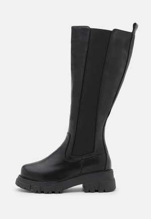 Black knee-high boot with chunky lug sole, elastic panel along the shaft, and pull tab at the back for easy wear.