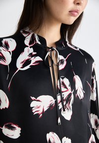 Svart bluse med hvit og rød blomsterdesign, kort mandarin krage, og en nøkkelhullåpning med knyte-detalj og metalliske accenter.