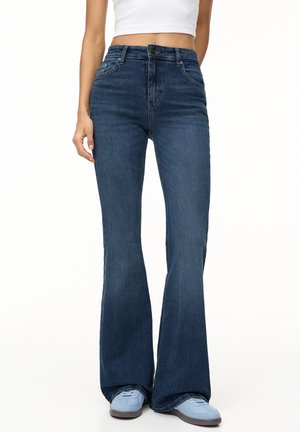 Femme portant un jean évasé taille haute bleu foncé, un crop top blanc et des baskets bleu clair, debout sur un fond blanc.