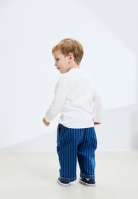Barn klädd i en vit tröja med långa ärmar och blå randiga byxor med relaxad passform, som vänder sig lite åt sidan.