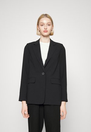 Blazer noir ajusté avec un bouton unique, revers crantés, deux poches avant et épaules structurées. Porté sur un haut blanc.