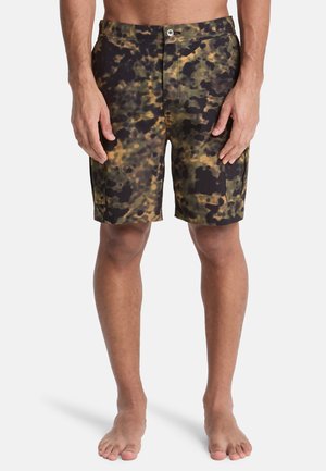 Mann trägt knielange Shorts mit grünem, schwarzem und braunem abstraktem Tarnmuster, steht barfuß vor weißem Hintergrund.