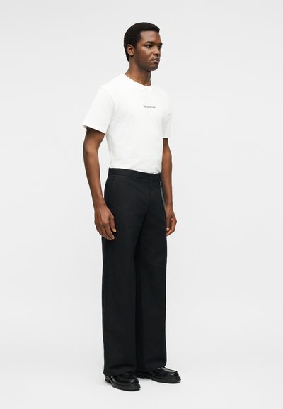 Άνδρας που στέκεται, φοράει λευκή κοντομάνικη μπλούζα, μαύρο φαρδύ παντελόνι, μαύρα παπούτσια, μπροστά από απλό φωτεινό γκρι φόντο.