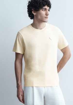 Ung mann med krøllete hår iført en lys beige t-skjorte og hvite bukser, stående med en hånd på hoften, ser mot høyre mot en ensfarget bakgrunn.