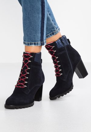 Granatowe zamszowe botki za kostkę na masywnym obcasie, z czerwono-białymi sznurówkami i bocznymi zamkami, noszone z krótkimi niebieskimi dżinsami.