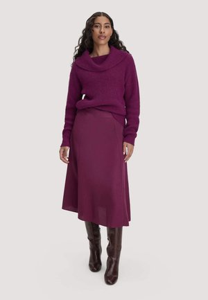 Frau mit langen lockigen Haaren, die einen violetten Rollkragenpullover, einen passenden violetten Midi-Rock und dunkelbraune kniehohe Stiefel trägt und nach vorne blickt.
