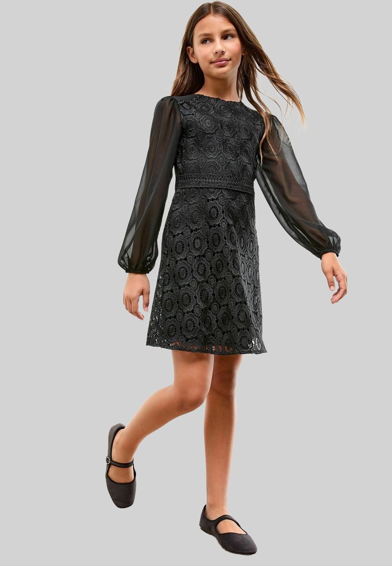 Fille marchant dans une robe en dentelle noire avec des manches longues transparentes et des chaussures Mary Jane noires, sur un fond gris uni.
