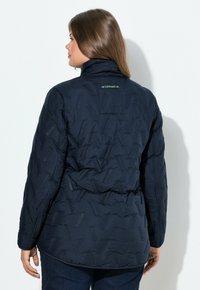 Mörkblå puffjacka med en texturerad yta, hög krage och en grön logotagg på baksidan. Bärs med mörka jeans och visar långa ärmar.