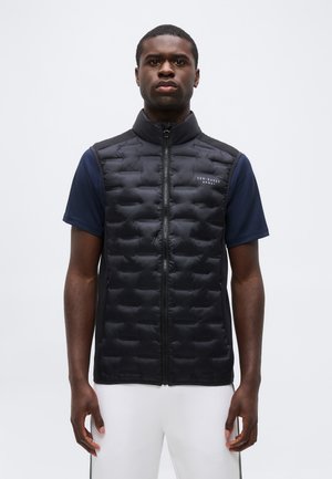 Mężczyzna w czarnej pikowanej kamizelce na granatowej koszuli z krótkim rękawem i białych spodniach, stojący na jasnym, jednolitym tle.