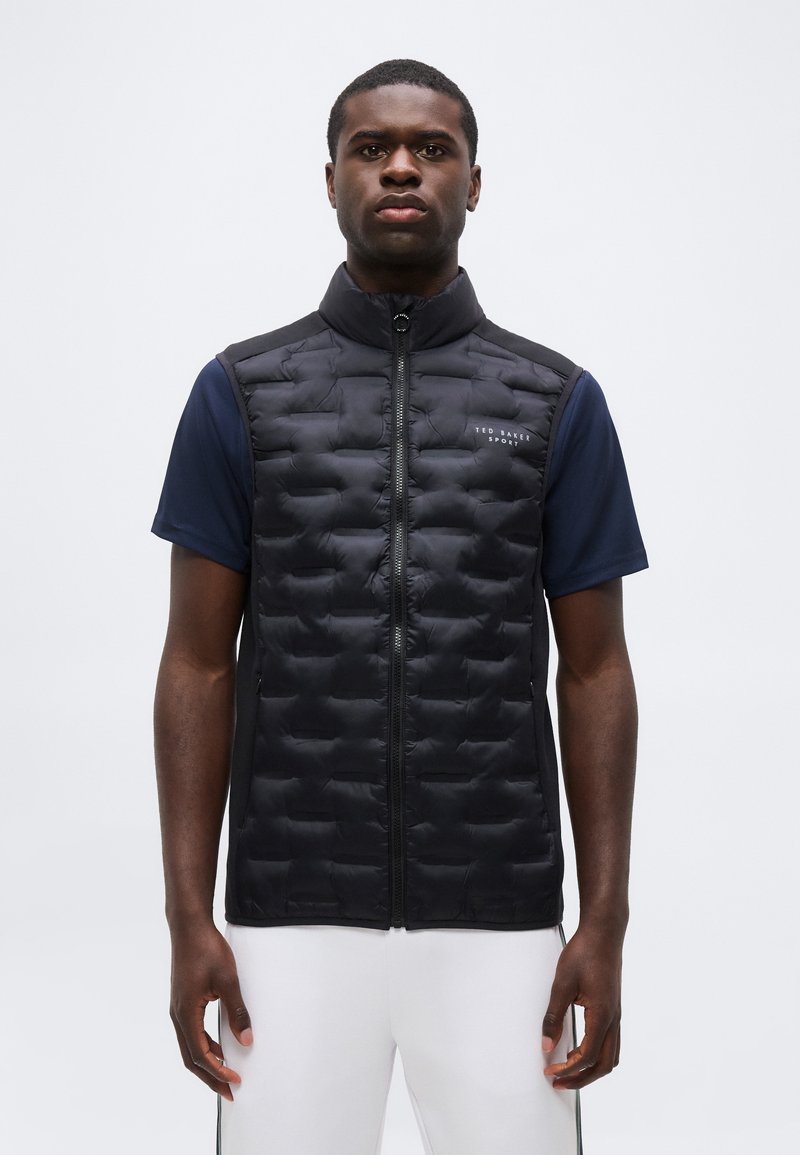 Άνδρας φοράει μαύρο αμάνικο καπιτονέ γιλέκο πάνω από μπλε σκουρόχρωμη κοντομάνικη μπλούζα και λευκό παντελόνι, στέκεται μπροστά σε απλό φωτεινό φόντο.