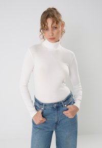 Bijela, dugosleeve majica s vunenim ovratnikom izrađena od glatkog materijala, u paru s trapericama svijetloplavih tonova visoka struka, s kopčanjem na dugme i patentni zatvarač.