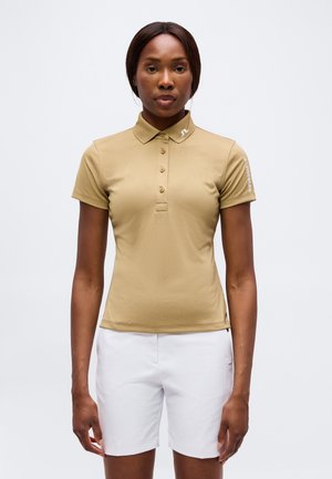 Kvinde iført en beige kortærmet polo og hvide shorts, stående stille mod en ensfarvet lys baggrund.