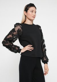 Sudadera negra con mangas de tela transparente decoradas con aplicaciones florales negras. Cuenta con un escote redondo y puños acanalados.
