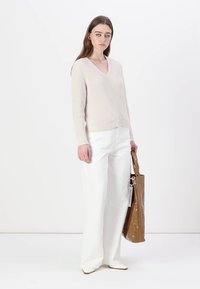 Pull en maille côtelée beige avec décolleté en V, associé à un pantalon large blanc et un sac fourre-tout brillant marron orné d'un accessoire en forme d'anneau en argent.