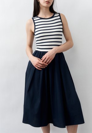 Vrouw draagt mouwloze jurk met marineblauw-wit horizontaal gestreept bovenstuk en effen marineblauwe A-lijn rok, staand met handen zachtjes gevouwen.