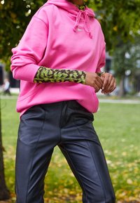 Pink hoodie with drawstrings and a Nike logo, layered over a green and black patterned long-sleeve top, paired with black leather pants.