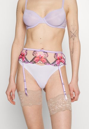 Kvinne iført en lilafarget bh, matchende strømpebelte med rosa sommerfuglbroderi, hvite truser og beige blondestøvler som går opp til lårene.