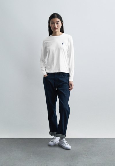 Νεαρή γυναίκα στέκεται με τα χέρια στις τσέπες, φορώντας λευκό μακρυμάνικο πουκάμισο, σκούρο τζιν με γυρισμένα μπατζάκια και λευκά αθλητικά παπούτσια, μπροστά σε απλό φόντο.